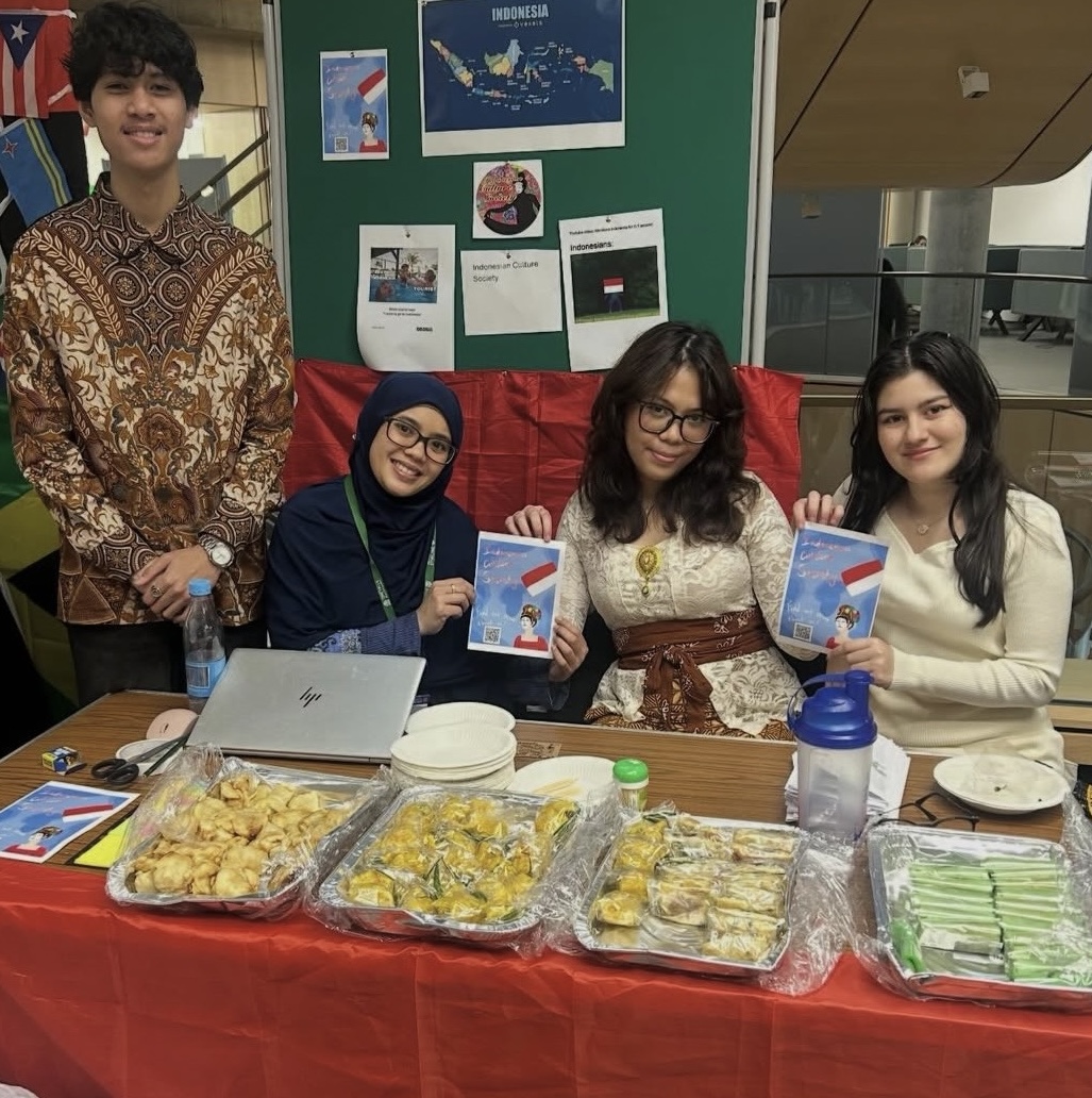 Group of students sat at a societies fair, they are holding up flyers to the camera with information about their society.