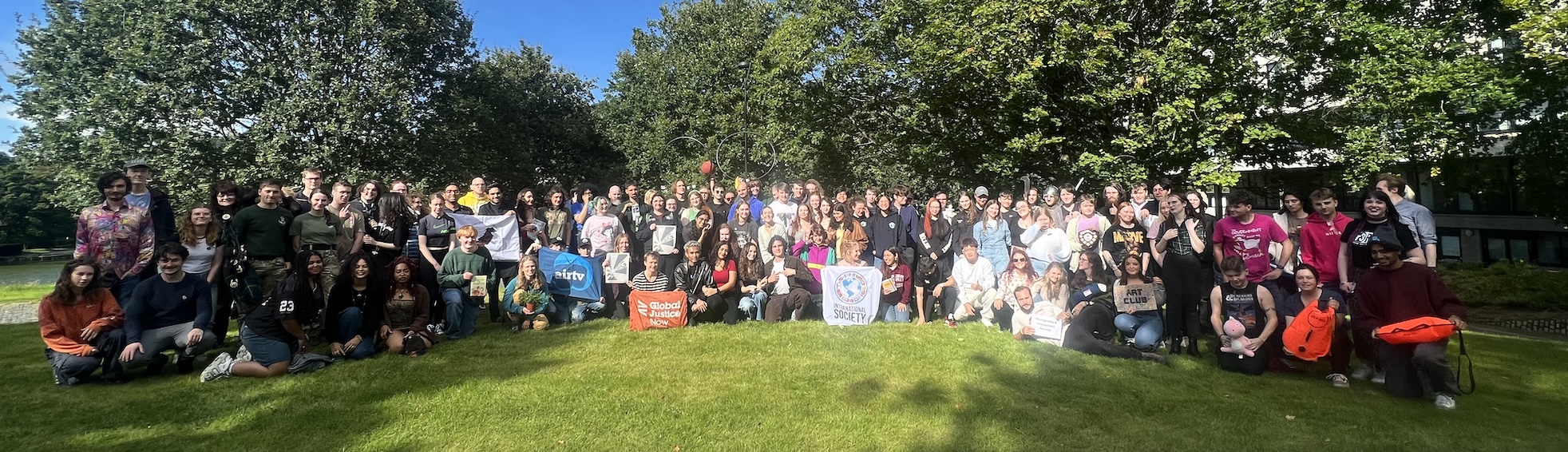 Group photo of around 100 students on a green lawn with a bright blue sky behind.