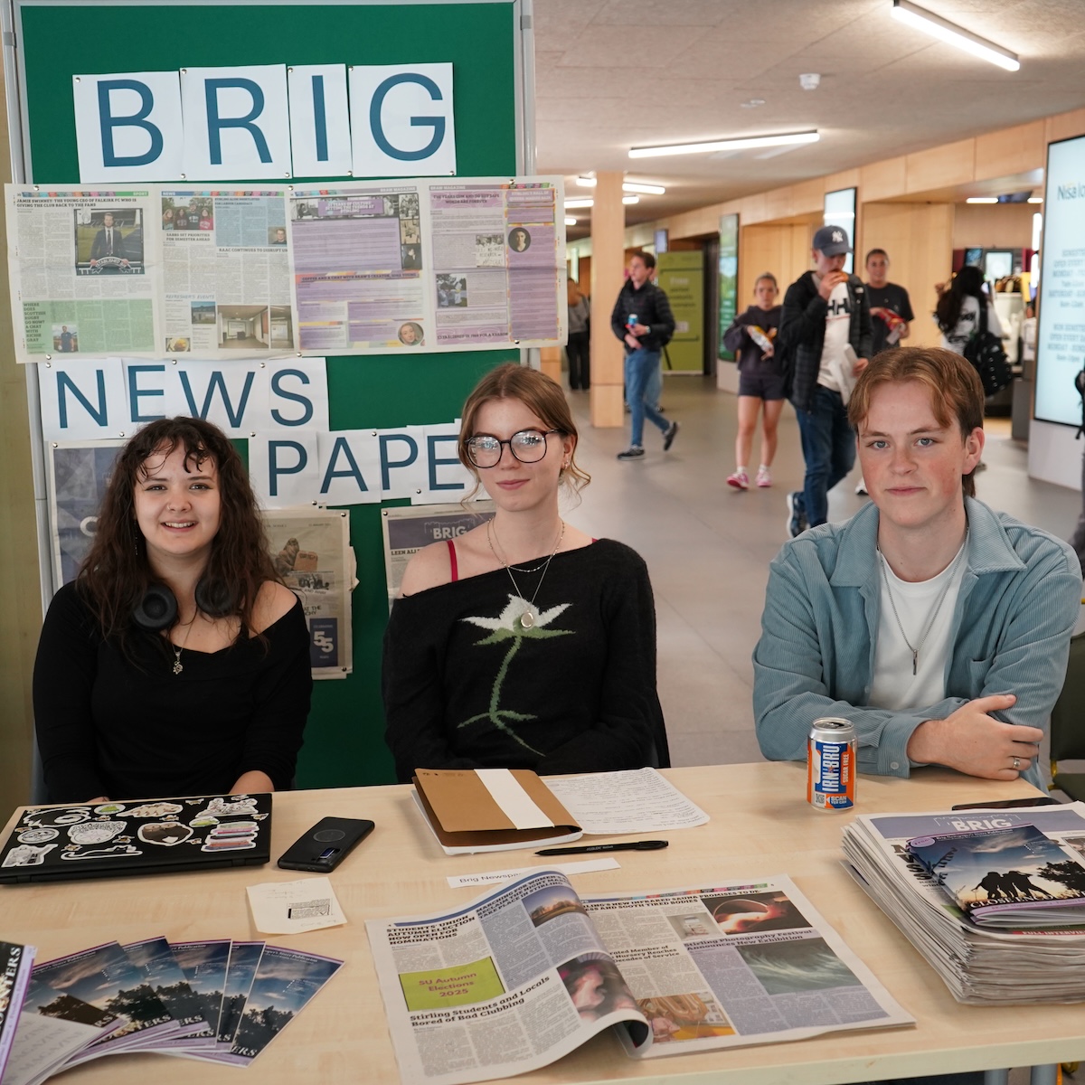 Three students sat behind a table with a sign behind reading Brig Newspaper