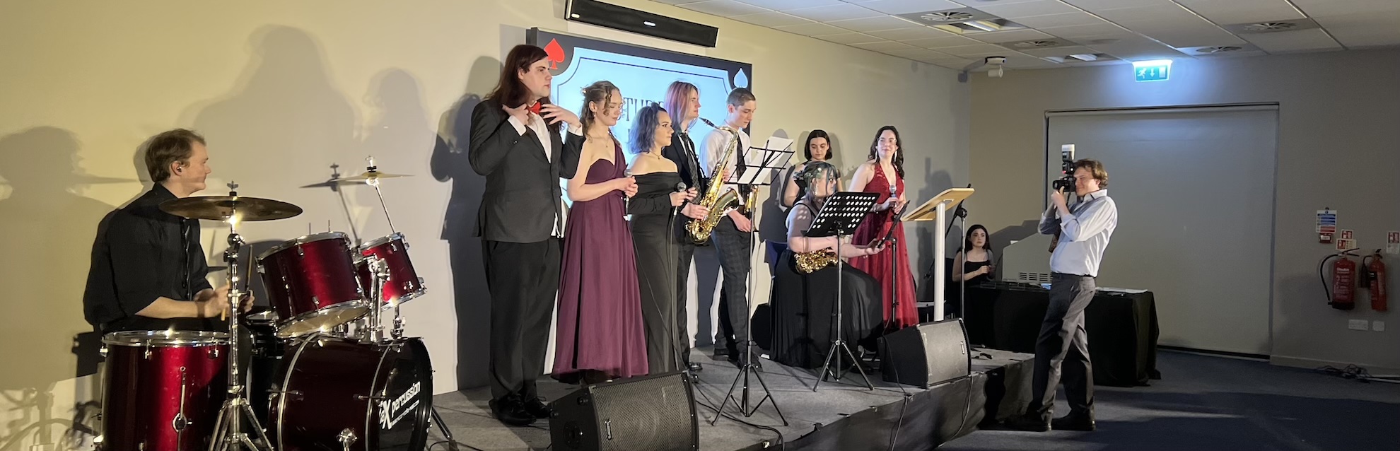 A 9 piece band on stage wearing smart dresses and suits playing inside a university room.