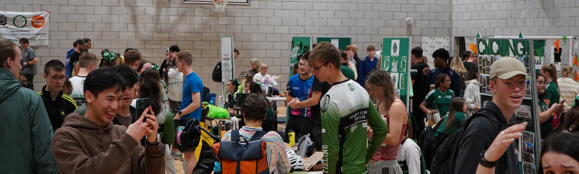 Busy sports hall filled with dozens of student stands, there is a person at the front holding up their phone smiling taking a photo on their phone.