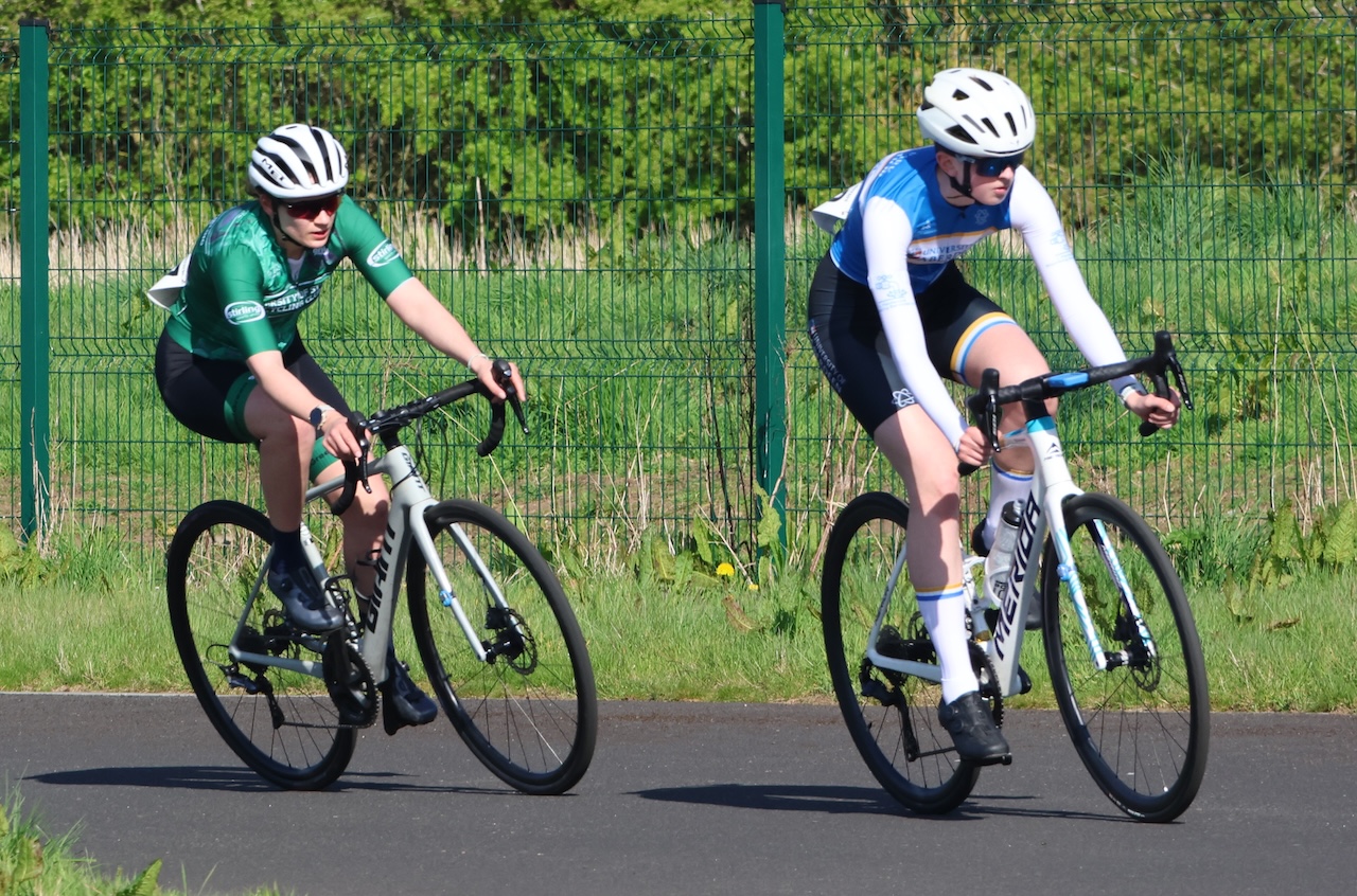 Two people on racing bikes wearing bike gear on a tarmac path with nature behind.