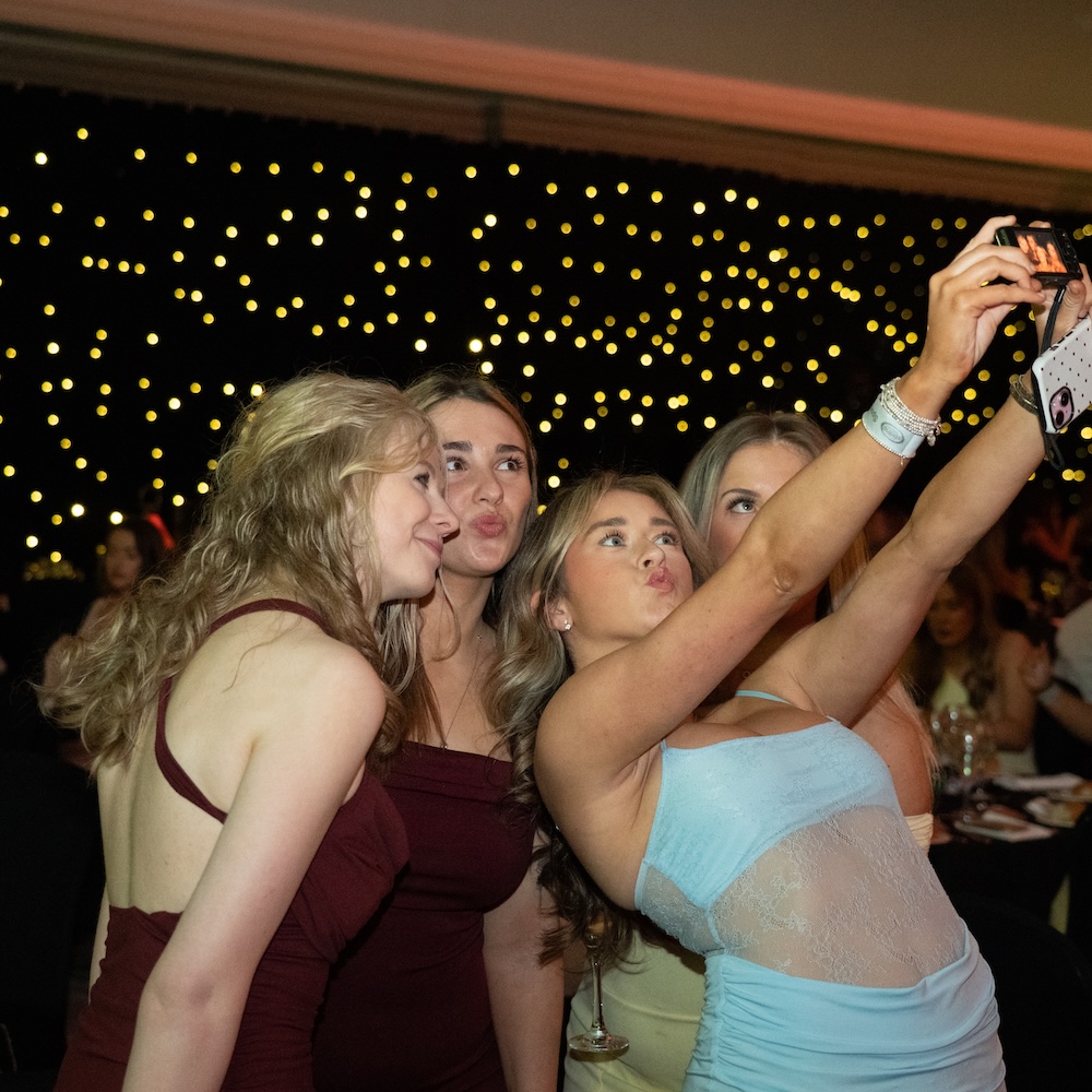 Group of girls wearing ball dresses, taking a selfie, there are fairy lights behind them.