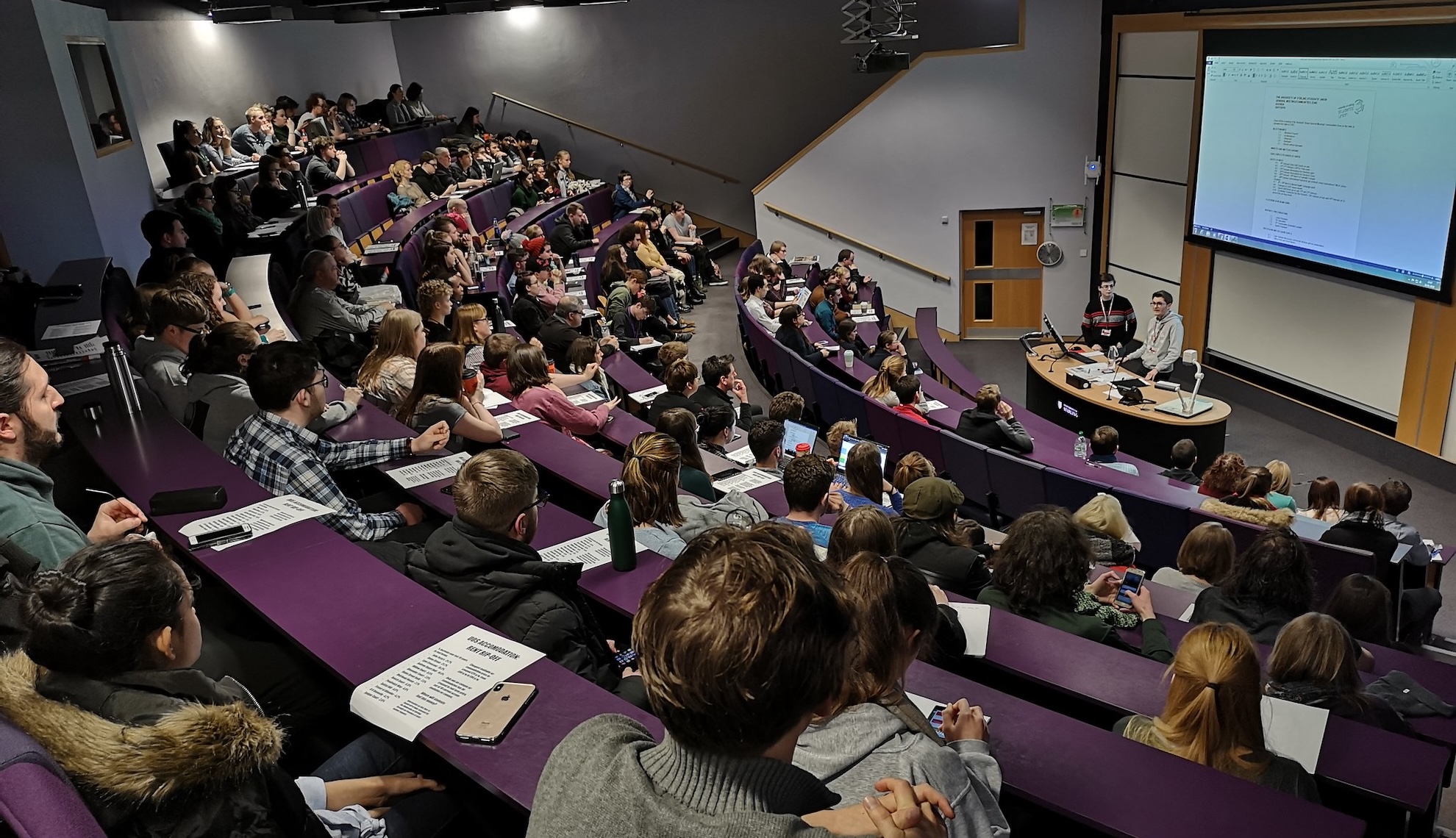 Large university classroom with many rows of seats and desks, looking down towards the screen and tutor at the front.