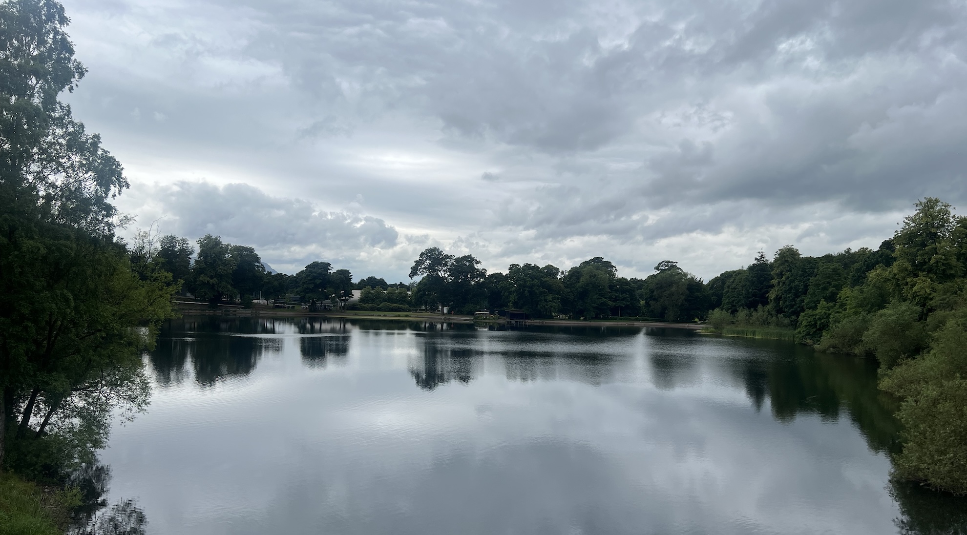 A large lake with trees in the background reflected against the water, the sky is grey and cloudy.