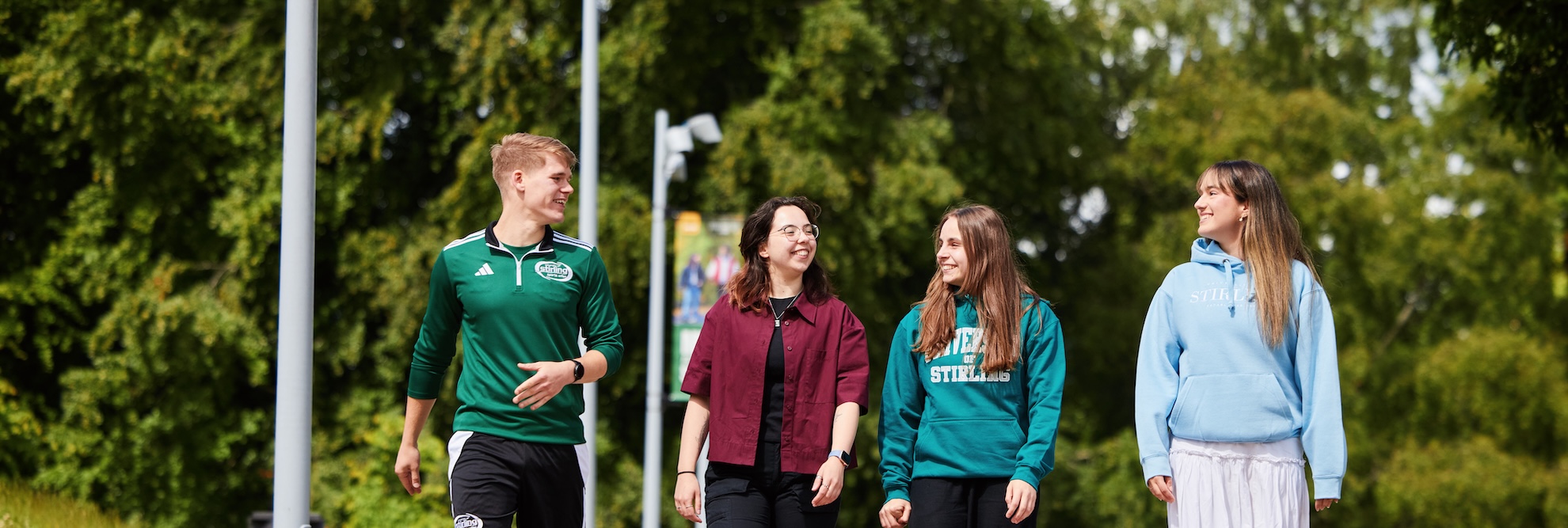 Four students walking along a path towards the camera in a university campus, lots of trees around and grass