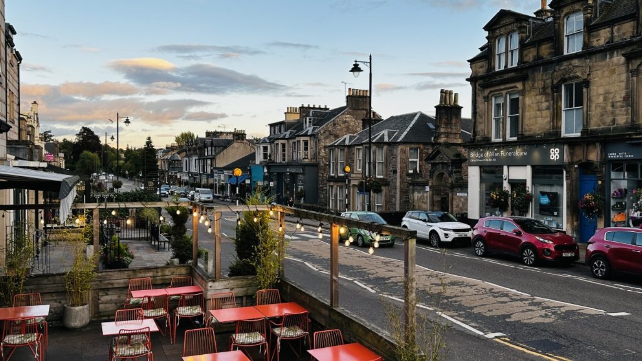 A street with a beer garden featuring red tables on the left and over the road there is a row of terraced houses