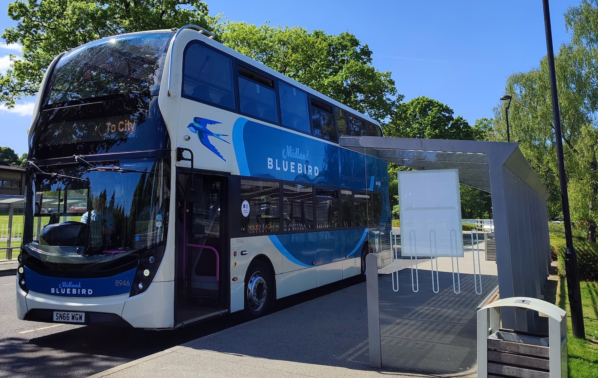 A white modern double decker bus stopped at a bus stop with trees behind and a bright blue sky.