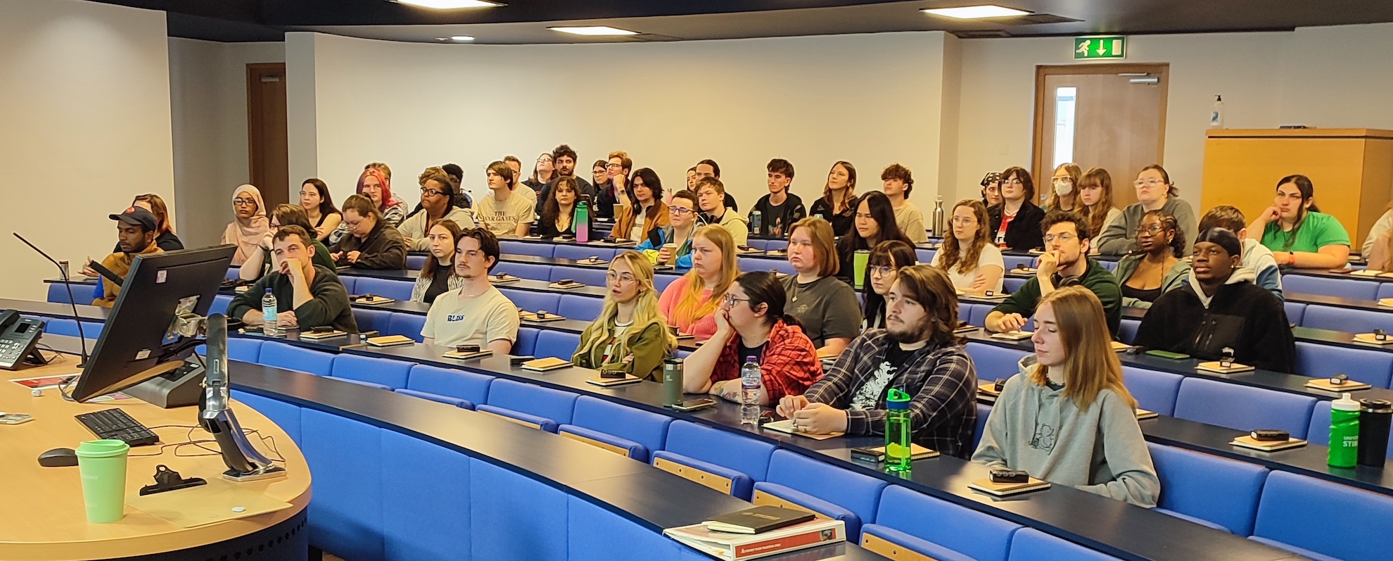 A lecture theatre with rows of blue seats, a class of students are looking towards the whiteboard.