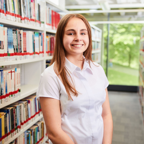 person with shoulder length light brown hair wearing a white shirt standing in a library