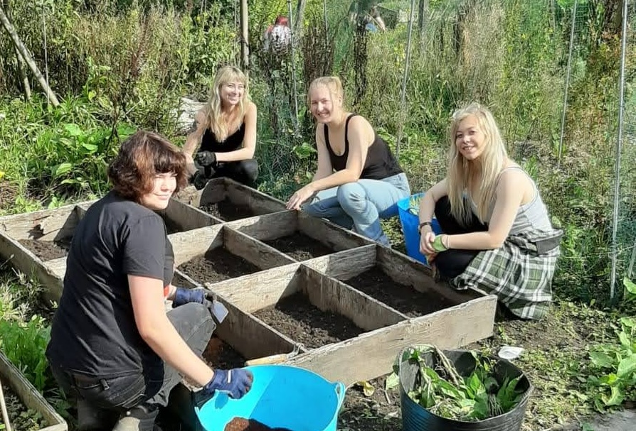 Four people working in a community garden, they are turned to face the camera while crouching down planting things in a flower bed.