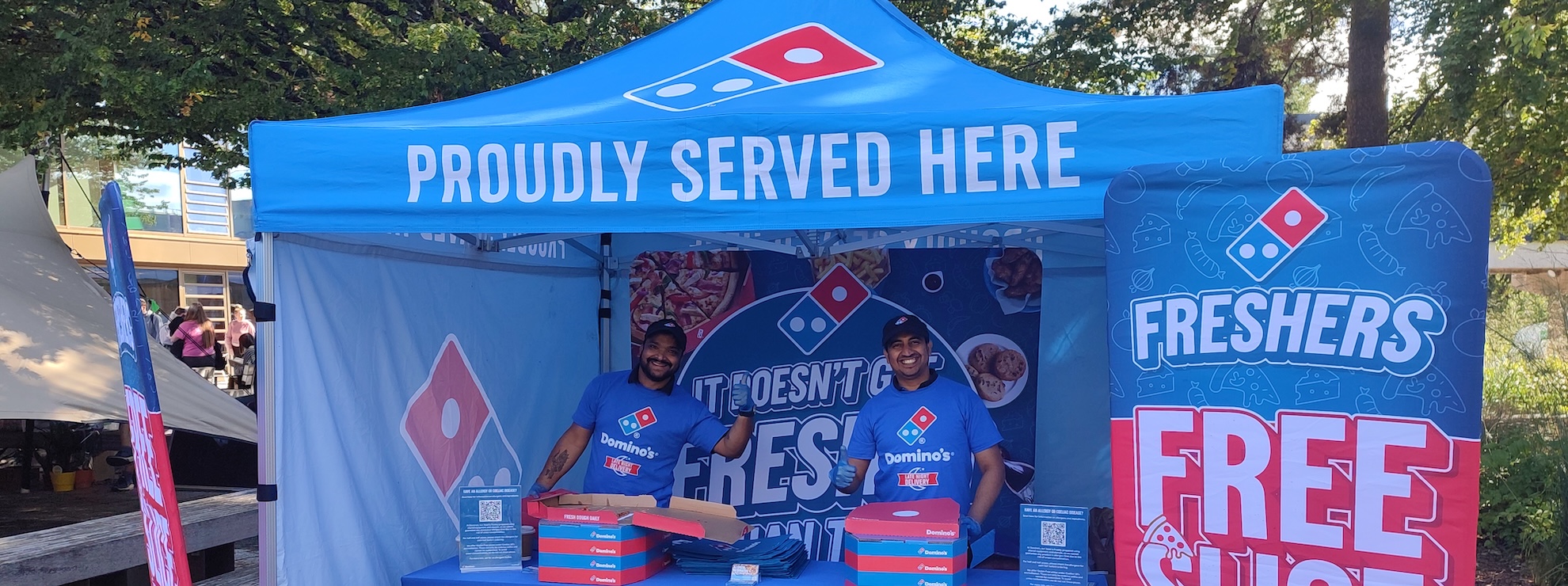A stall outdoors with bright blue and red canopy, two people are stood behind a table offering free pizza slices.