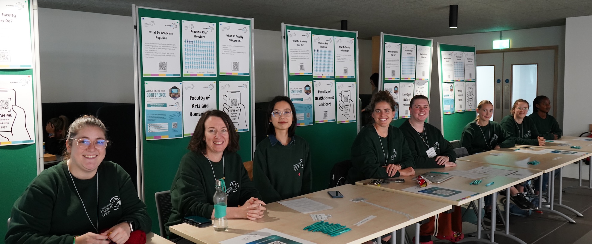 Five people sat behind a table at an academic representatives fair, they have a board behind them with information about reps.